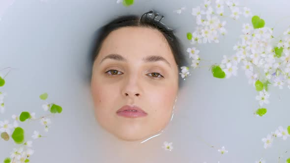 Brunette Girl Lies in a White Bathtub with Milk and Small Flowers Floating