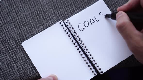 A male hand writes GOALS in a notebook on a gray textural surface of a table