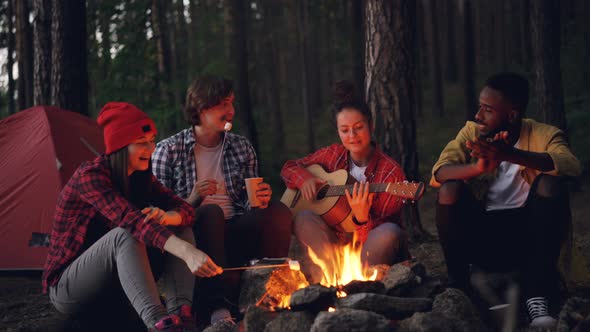 Happy Adventurous Hikers Are Sitting Around Fire, Singing and Playing the Guitar, Eating Marshmallow