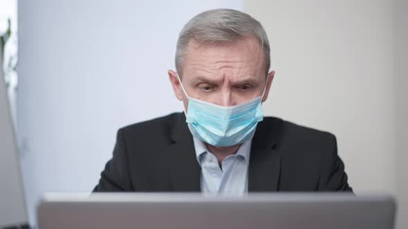 Portrait of Worried Caucasian Senior Man in Covid19 Face Mask Messaging Online on Laptop Sitting in