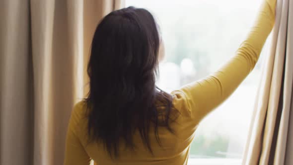 Back view of biracial woman revealing curtains and looking outside window