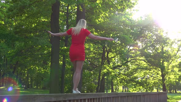 A Young Beautiful Woman in a Red Dress Walks Along a Wooden Railing in a Park on a Sunny Day