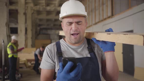 Close-up Portrait of Cheerful Caucasian Guy in Protective Helmet Looking at His Smart Phone and