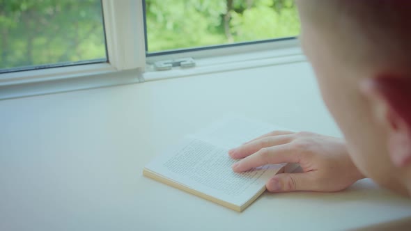 Young Man Reading a Book Sitting at an Open Window on a Sunny Day Back View