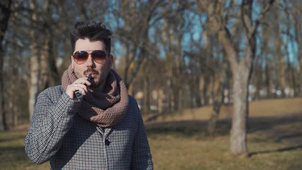 A Young Handsome Bearded Hipster Man Vaping an Electronic Cigarette in the Park in the Spring. Close