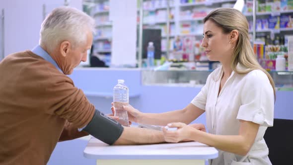 Side View Smiling Caring Female Pharmacist Passing Water for Unwell Senior Man Taking Off Blood