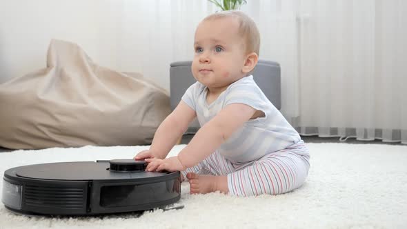 Happy Smiling Baby Boy Sitting on Carpet and Playing with Robot Vacuum Cleaner
