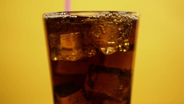 Mixing Cold Cola Cocktail with Pink Drinking Straw. Glass Full of Fizzy Drink Ice Cubes on Yellow BG