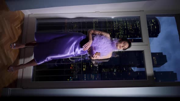 Rich Woman in a Purple Dress Standing By the Window with a Glass of Champagne and Looks at the Night