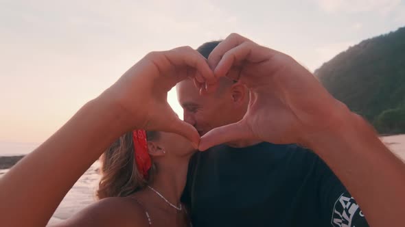 Happy Young Caucasian Man and Woman Makes Heart Shape and Gently Kisses on Beach