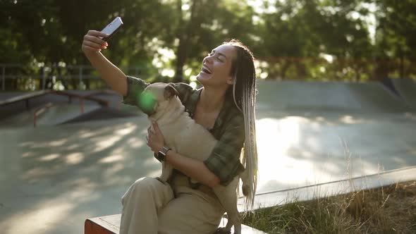 Cheerful Girl Taking Selfie Photo with Cute Pug Puppy in Green City Park Holding Smartphone in