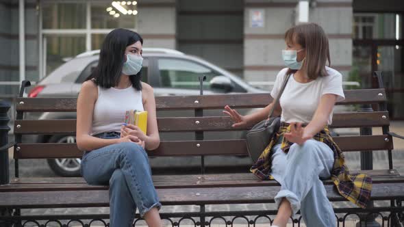 Wide Shot of Two Positive Teenage Girls in Covid Face Masks Sitting on Bench with Backpacks and