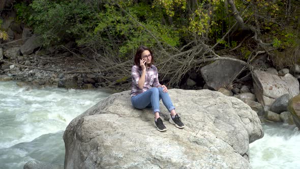 A Young Woman Sits on a Stone and Speaks on the Phone in the Mountains By the River.