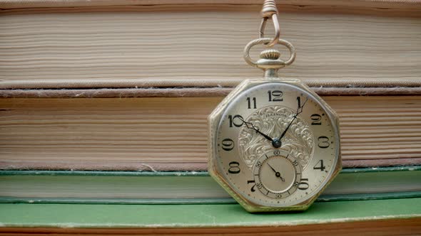 Antique Gold Pocket Watch on Chain Hanging Against Background of Stacks of Books