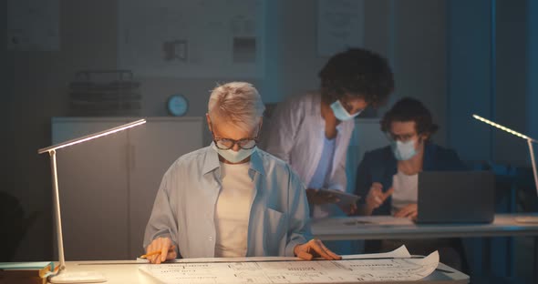 Portrait of Mature Woman in Protective Mask Working with Blueprints in Dark Office