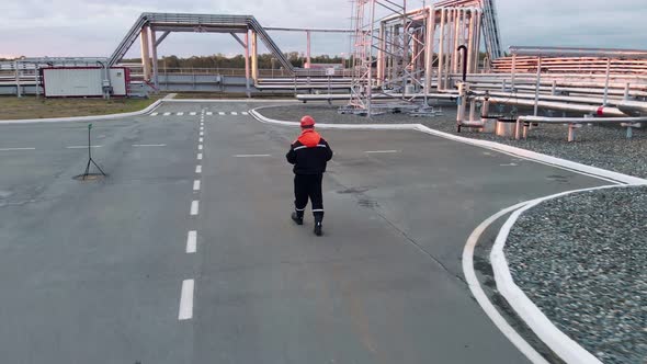 A Drone Flies Around a Worker in an Industrial Area Who Bypasses an Area with an Oil and Gas