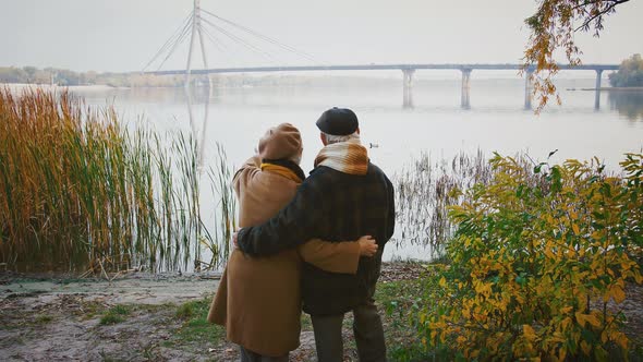 Loving Aged Couple Hugging and Smiling Standing on Bank of a River and Looking at Huge Bridge