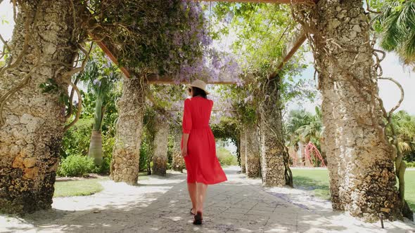 Young Brunette Woman in Red Dress Walks Through the Garden and Turns Around Smiling