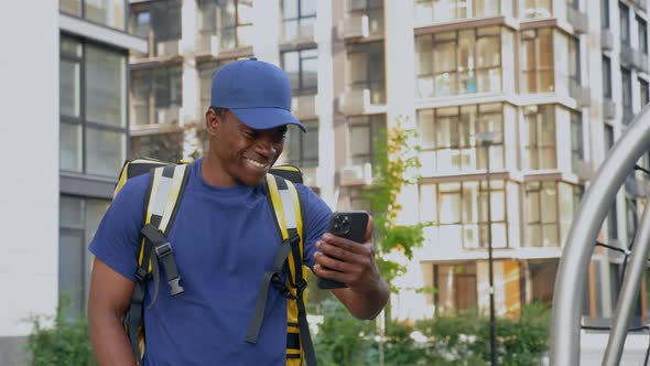 Smiling African Man Courier Food Delivery Walks Street Watching in Smartphone