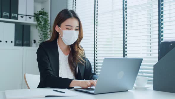 Asian businesswoman in formal suit use laptop computer wear facemask and work at workplace.