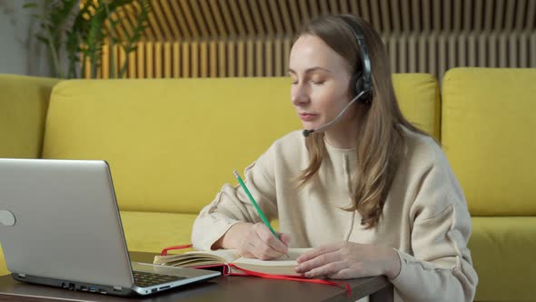 Woman in Headset Sitting on Yellow Couch at Home and Video Chatting on Laptop Computer