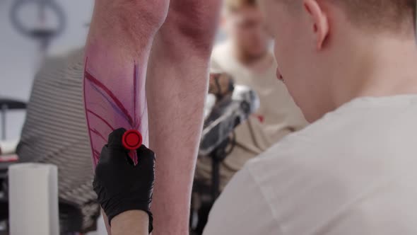 Young Man Tattoo Artist Drawing a Sketch on the Leg on His Client with a Red Marker