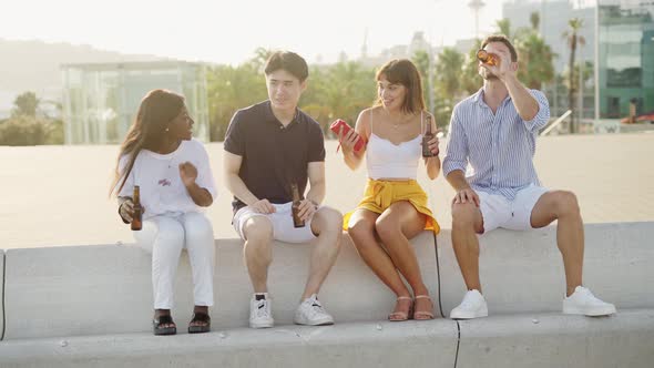 Four Multiracial People Best Friends Listening to Music and Drinking Beer on Embankment at Sunset