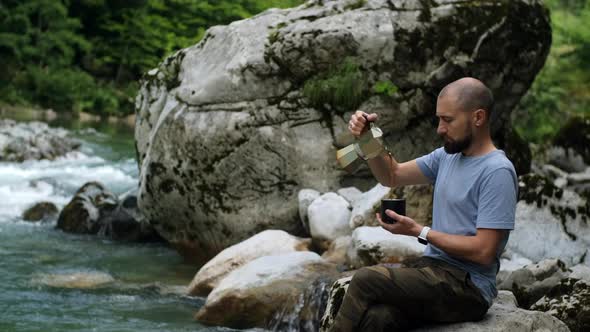 Traveler man pours coffee  from Mocha Pot against mountain river, road trip