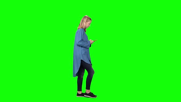 Blonde Teenager Girl Calmly Walking and Reading Text Message on Mobile Phone at Greenscreen. Profile