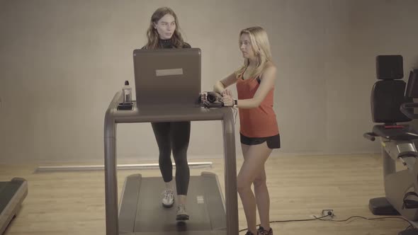 Blond Personal Trainer Adjusting Speed on Treadmill and Talking To Young Brunette Caucasian Woman