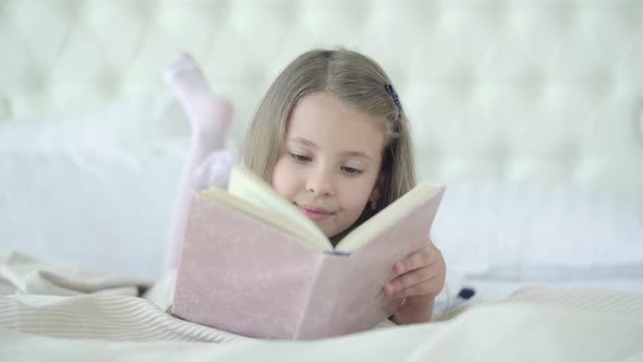 Pretty Brunette Caucasian Girl Lying on Bed Turning Book Pages and Smiling. Portrait of Relaxed