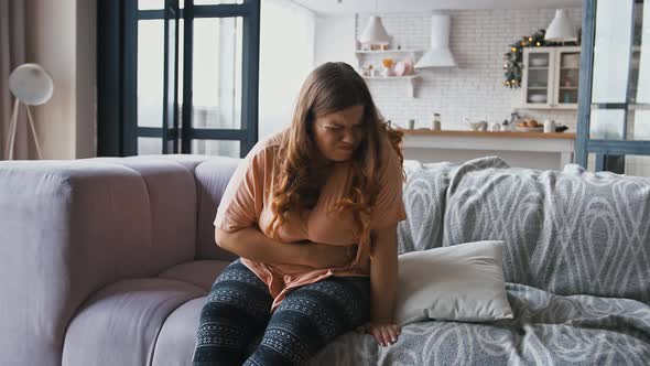 Young Overweight Woman Suffering From Abdominal Pain Period Cramps Sitting on Couch at Home