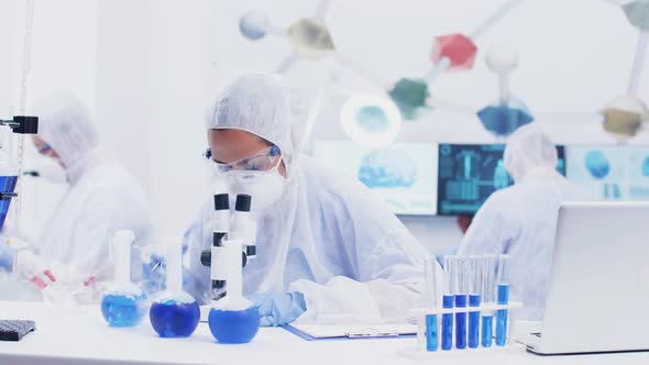 Female Scientist in Coverall Equipment Writing Notest and Looking Through Microscope