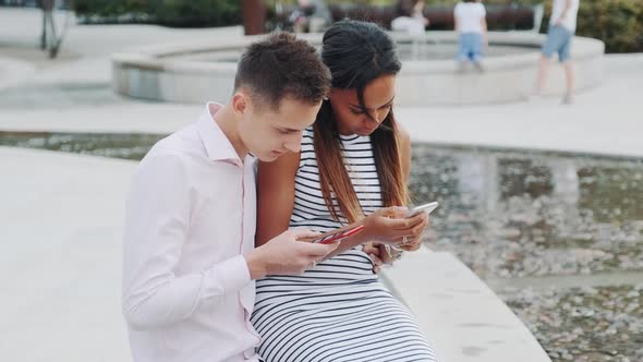 Multiethnic Couple Sitting Together Outdoors and Looking in the Smartphones