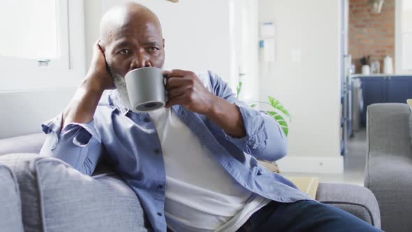 Thoughtful senior african american man in living room sitting on sofa, drinking coffee