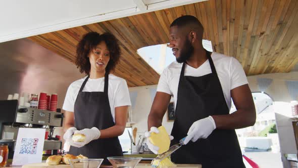 African american couple wearing aprons smiling while preparing hot dogs together in the food truck