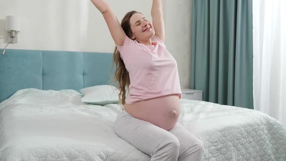 Happy Smiling Pregnant Woman in Pajamas Waking Up in Morning and Looking Out of the Window