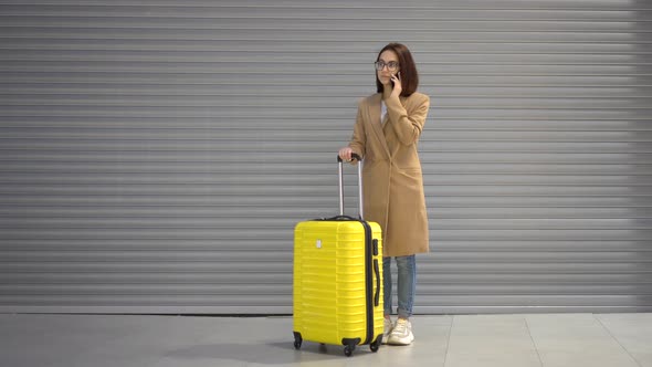 A Young Woman Stands with a Suitcase and Speaks on the Phone. Girl in a Coat on a Gray Background.