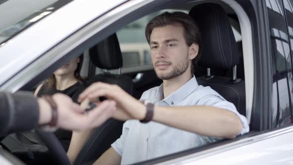 Portrait of Confident Young Caucasian Man Receiving Car Keys From Dealer in Dealership. Handsome