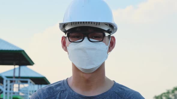 Young Asian engineer wearing a helmet and mask looks and smiles at the camera on the dam background.