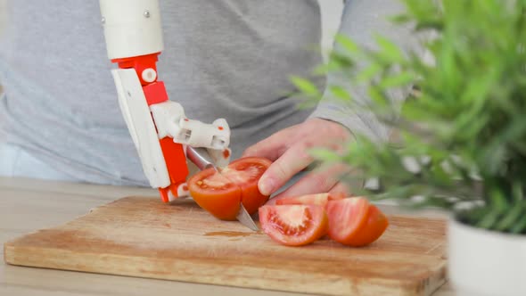 Man with Robotic Prosthetics Hand with Knife on It Is Trying To Cut Tomato on Wooden Board