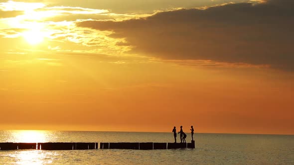 Four silhouettes of teenage children jump into the sea from breakwaters at an orange sunset