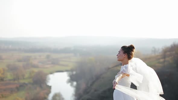 Happy Pretty Bride in Chic White Wedding Dress Is Standing in Deserted Field on Windy Day and Waving