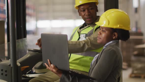 African american male and female workers wearing safety suits and using laptop in warehouse