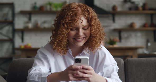 Smiling Redhead Young Woman Holding Modern Smartphone Texting Message at Sofa at Home