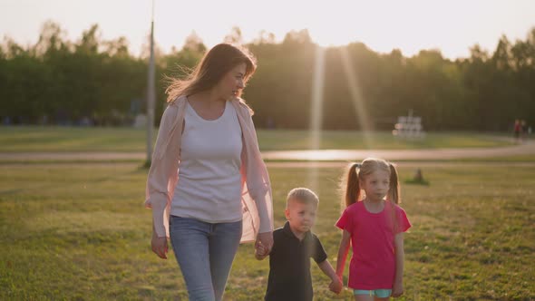 Happy Woman and Little Kids Walk Along City Park in Evening