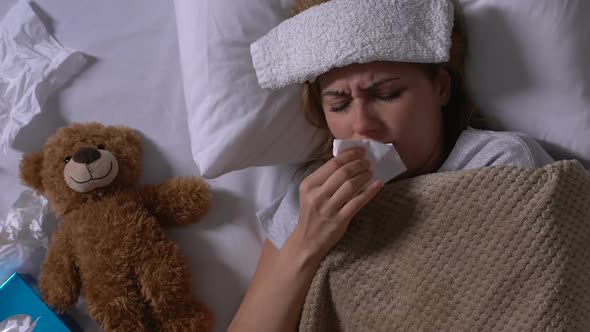 Ill Woman With Towel on Forehead and Runny Nose Hugging Teddy Bear in Bed
