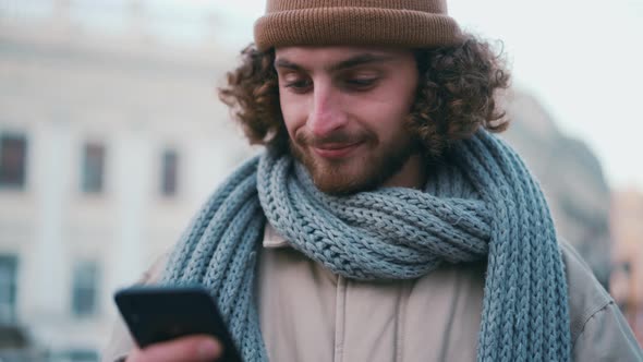Positive curly-haired man texting by phone