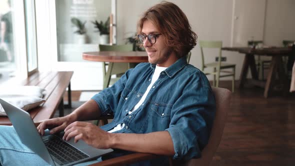 Laughing blond man in eyeglasses typing on laptop