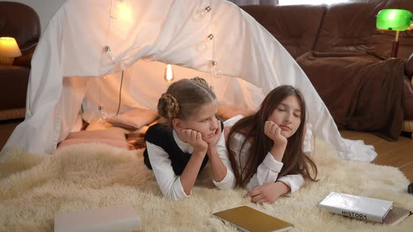 Two Charming Teen Girls Lying at Tent in Living Room with Books Talking